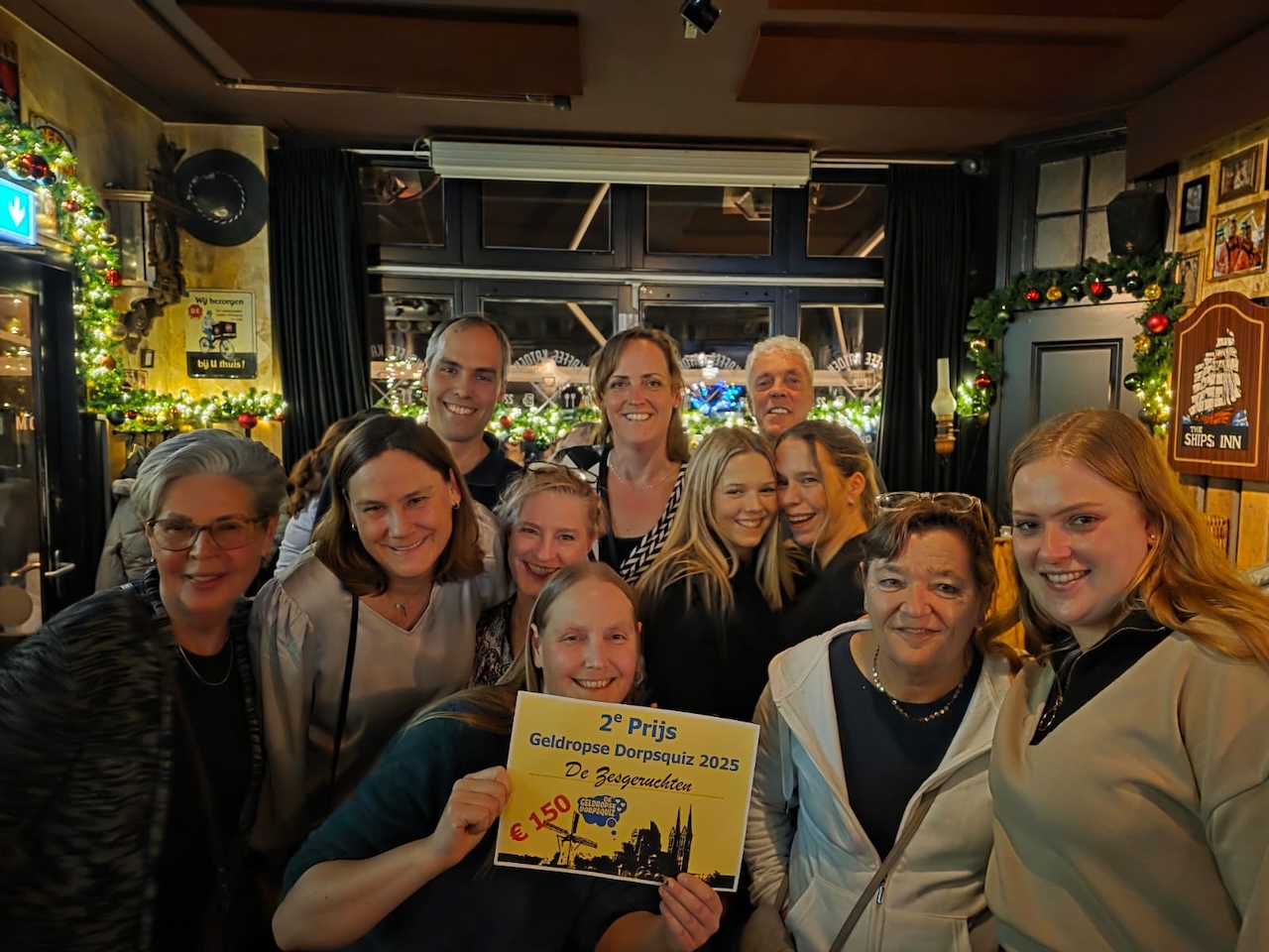 Groep mensen viert winst tweede prijs in dorpsquiz, binnen kerstdecoratie.