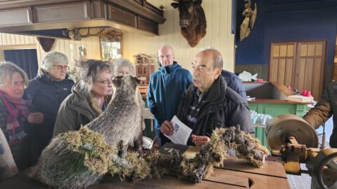 Groep mensen luistert naar een man die iets uitlegt met een dassenhuid en stam in een interieur.