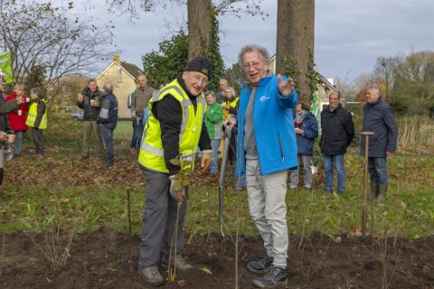 Mensen planten bomen in een park; twee mannen lachen terwijl ze een schep vasthouden.