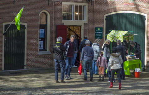 Groep mensen bij een marktstalletje voor een bakstenen gebouw met groene deuren.