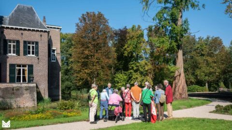Een groep mensen staat bij een historisch gebouw in een groene tuin op een zonnige dag.
