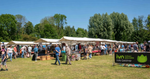 Buitenmarkt met kraampjes en mensen, omgeven door groene bomen. IVN Geldrop bord zichtbaar.