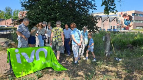 Groep mensen verzameld rond een tafel met een groene "IVN beleef natuur" banner buiten.