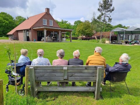 Ouderen zitten op een bank in een tuin, voor een huis met een glazen kas ernaast.
