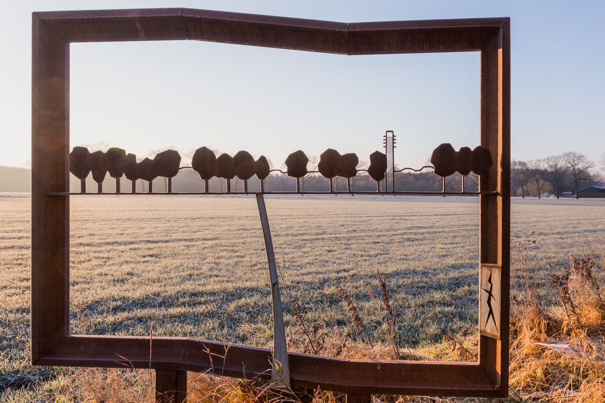 Roestige metalen sculptuur omlijst een rij bomen in een open, licht besneeuwd veld bij zonsopgang.