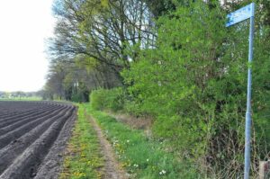 Landweg langs akker met groene struiken en bomen, bord met straatnaam aan rechterkant.