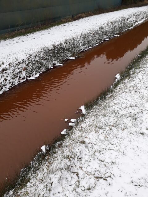 Bruine sloot met besneeuwde oevers en gras, onder bewolkte lucht.