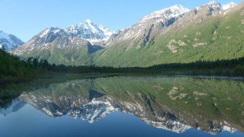 Bergen met besneeuwde toppen weerspiegeld in een kalm meer, omringd door groene bossen.