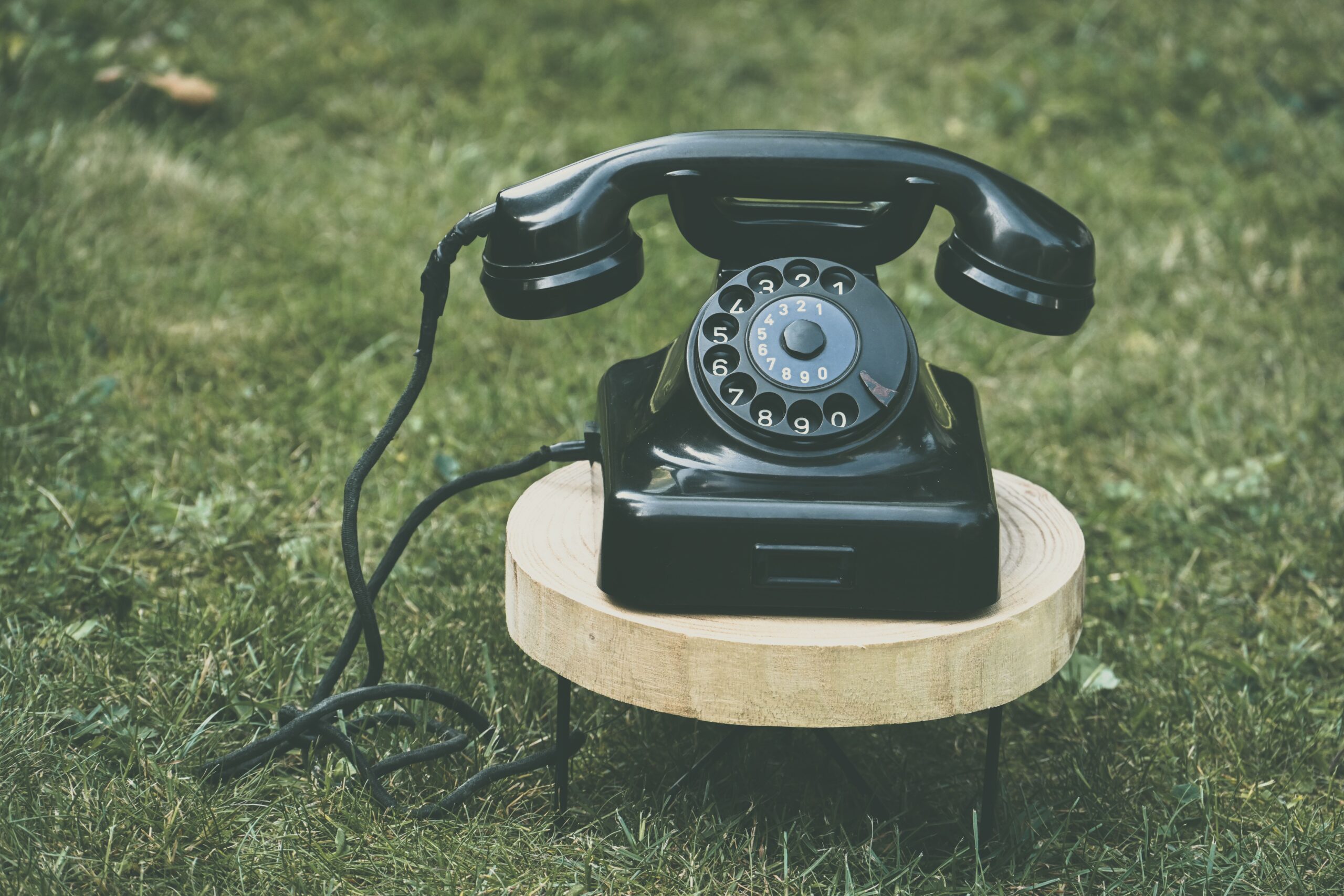 Zwarte draaischijftelefoon op houten krukje op gras.