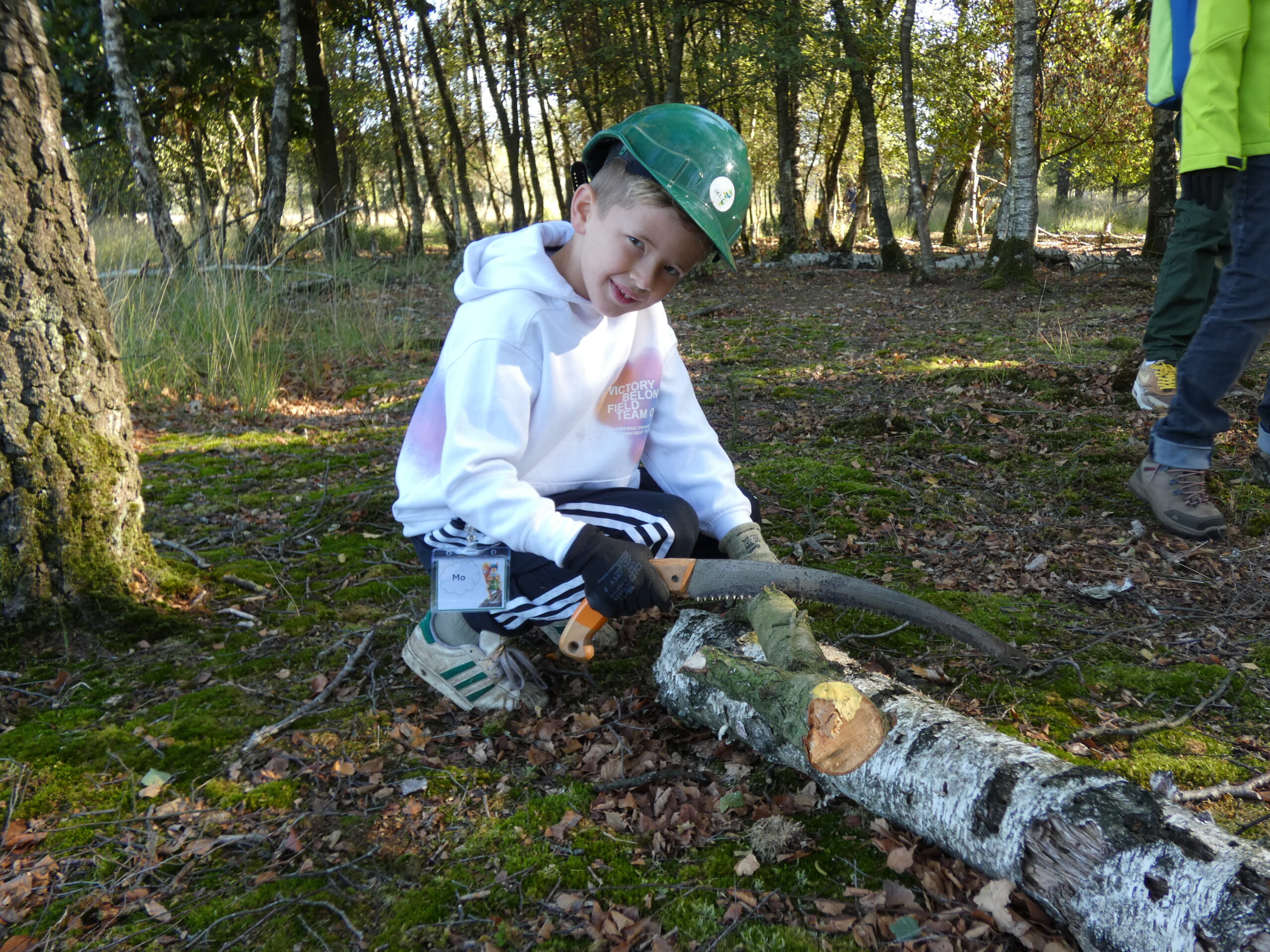Kind zaagt boomstam met handzaag in bos, draagt helm en handschoenen.