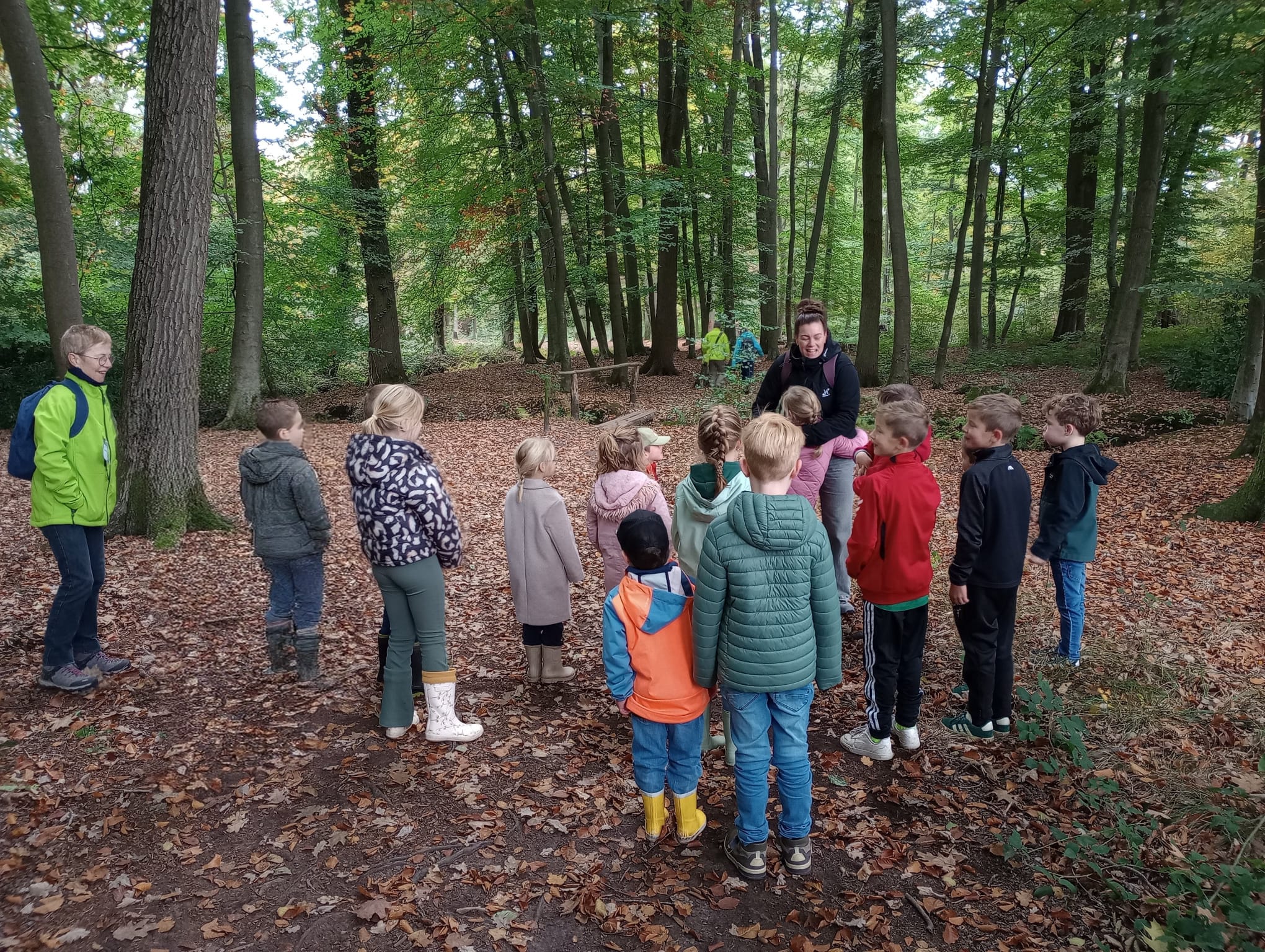 Groep kinderen en volwassene in herfstbos, omringd door bomen en bladeren op de grond.