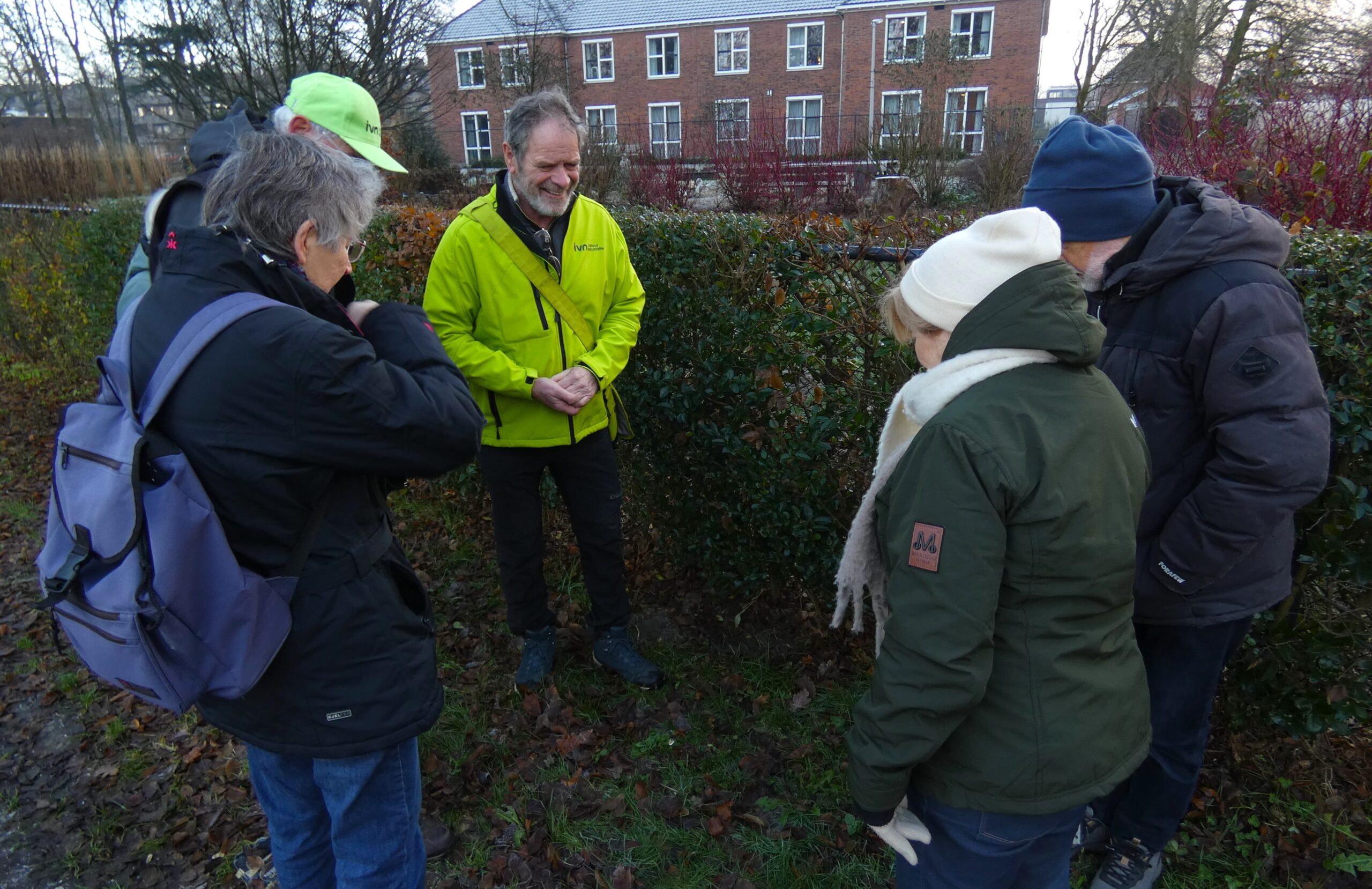 Een groep mensen staat buiten in een kring rond een gids in een gele jas, bij een heg.