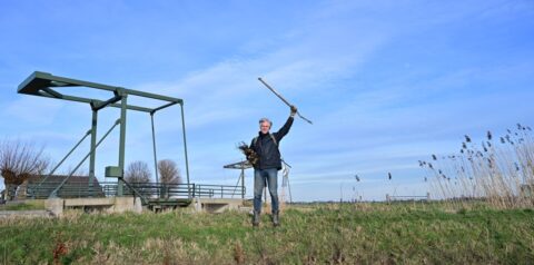 Persoon bij een brug op een grasveld, met een stok in de lucht gestoken.