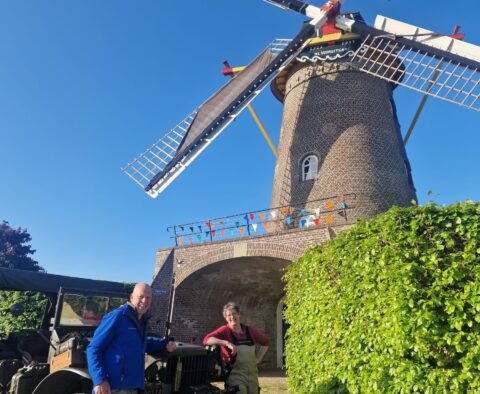 Twee mensen poseseren bij een klassieke auto voor een bakstenen windmolen met feestelijke vlaggetjes.