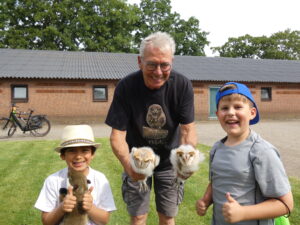 Een man en twee kinderen poseren met uilen, in de buitenlucht bij een bakstenen gebouw.