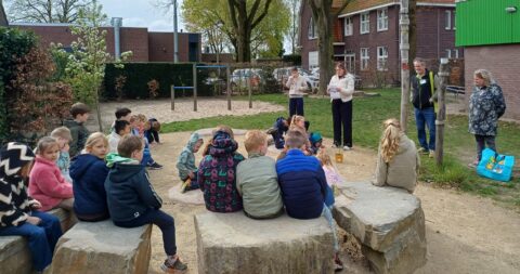 Groep kinderen luistert naar volwassenen in een schoolpleinsetting, met bomen en gebouwen op de achtergrond.
