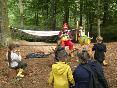 Een clown entertaint kinderen met een poppenshow in een bosrijke omgeving.