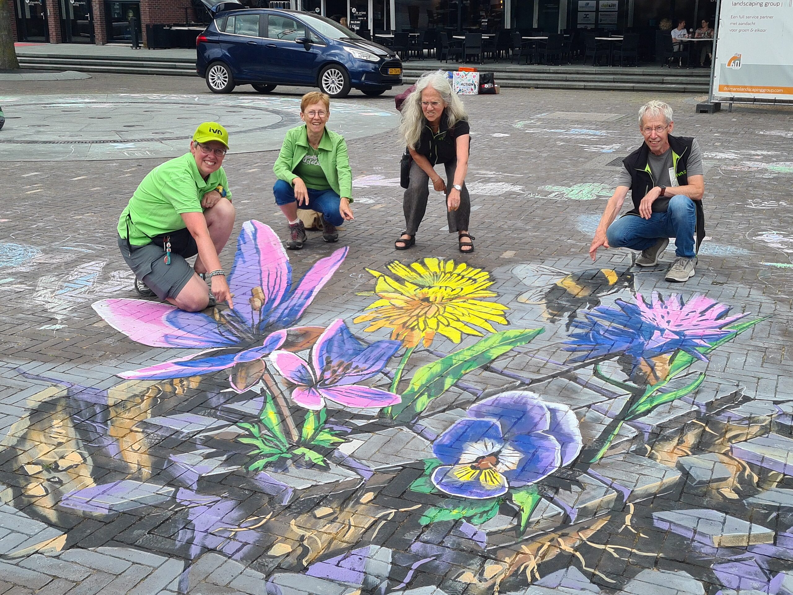 Vier mensen poseren rond kleurrijke krijttekening van bloemen en een bij op straat.