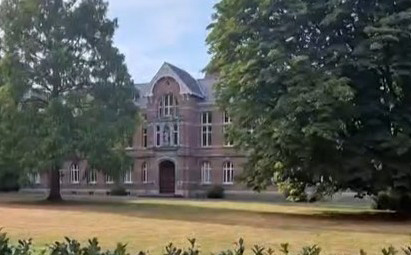 Oud bakstenen gebouw met puntdak, omgeven door bomen en grasveld.