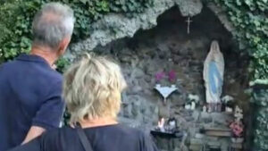 Een man en vrouw kijken naar een Mariabeeld in een grotaltaar met bloemen en kaarsen.