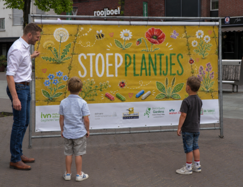 Man en twee kinderen bekijken kleurrijke poster over 'stoepplantjes' op een plein.