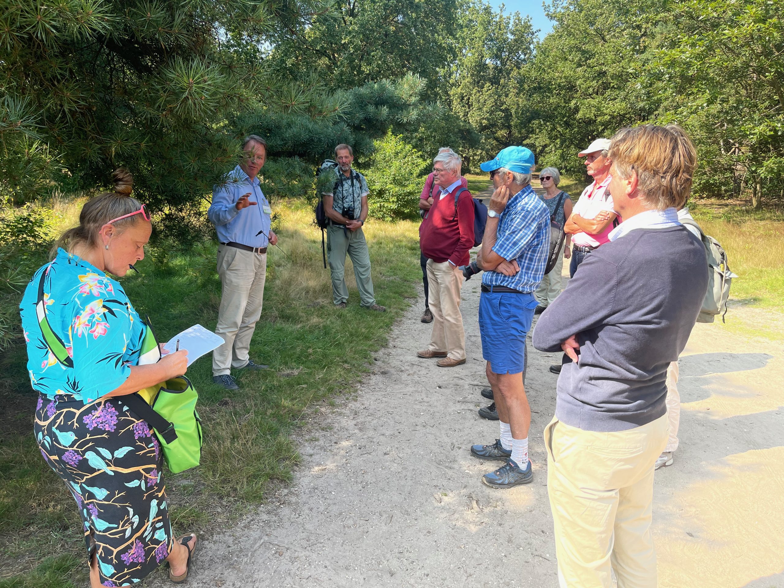 Een groep mensen luistert naar een spreker in een bosrijke omgeving. Eén persoon maakt notities.