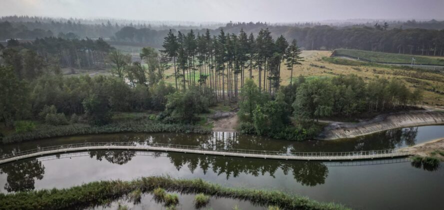 Luchtfoto van een kronkelende loopbrug over een rustige rivier met bomen en weelderig groen eromheen.