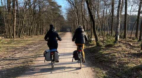 Twee mensen fietsen op een bospad, omgeven door kale bomen in de winterzon.