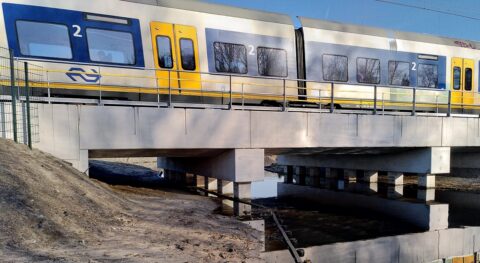 NS-trein op een brug, met blauwe en gele kleuren, weerspiegeld in het water eronder.
