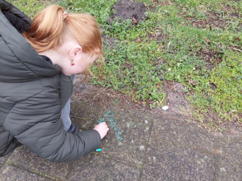 Kind schrijft met krijt op stoep naast gras. Ze draagt een groene jas.