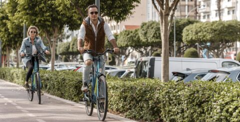 Twee mensen fietsen op een zonnig fietspad omgeven door bomen en geparkeerde auto's.