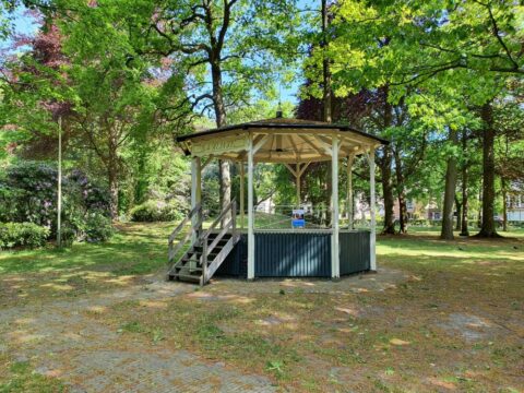 Houten prieel in een park met omringende groene bomen en bloeiende struiken.