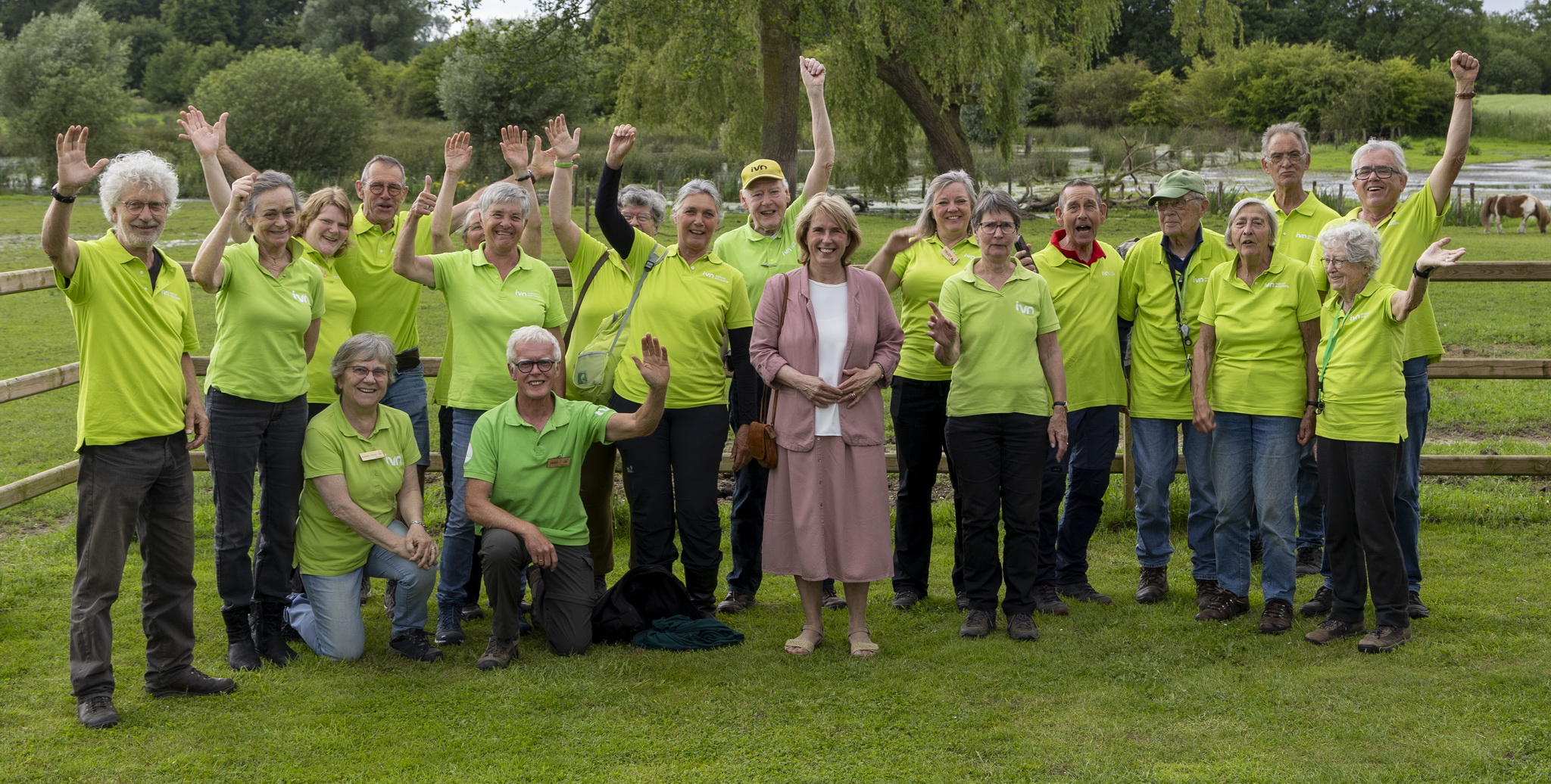 Groep mensen in groene shirts, buiten in een park. Sommigen zwaaien en staan in het gras.