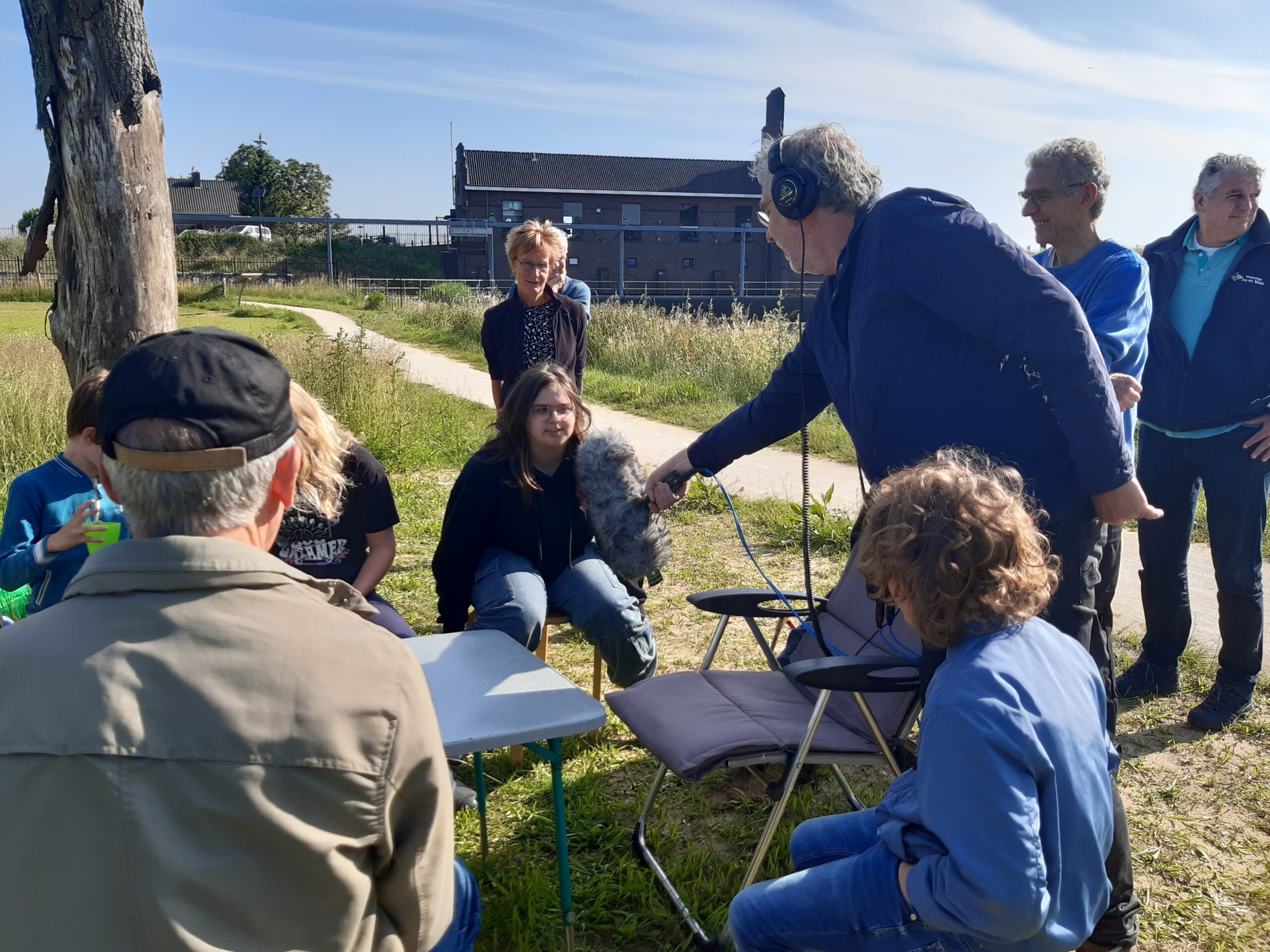 Groep mensen buiten, man met koptelefoon en microfoon spreekt met zittende jongere.