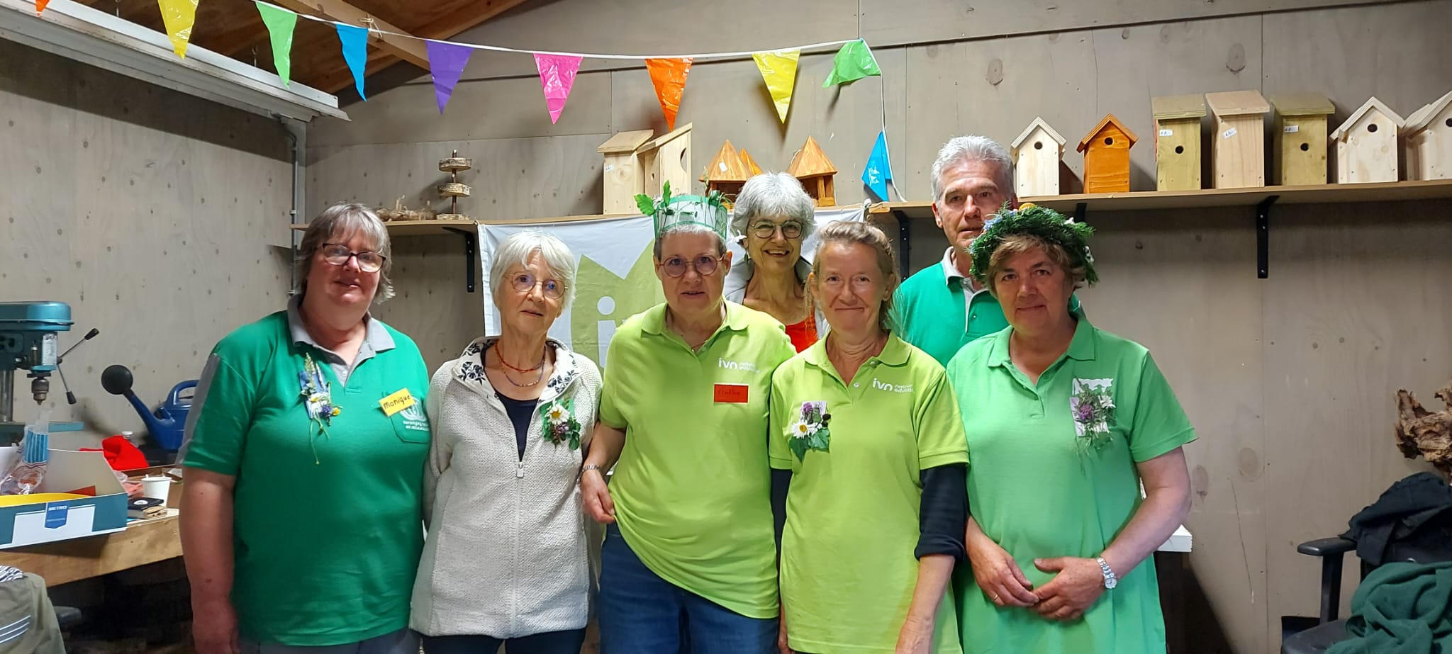 Groep mensen in groene shirts poseert in een werkplaats met vogelhuisjes op de achtergrond.