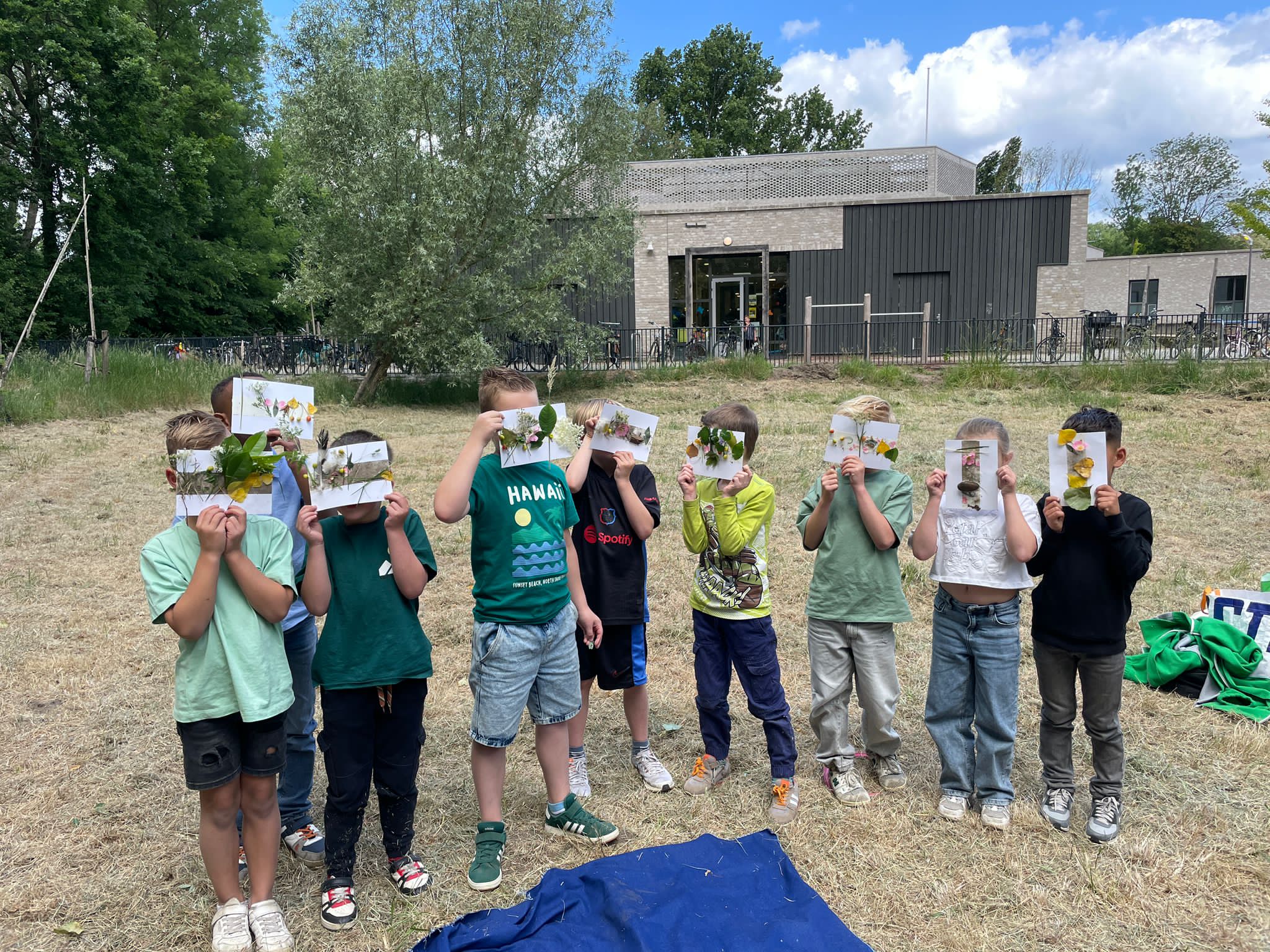 Een groep kinderen houdt tekeningen met bladeren vast voor hun gezichten, buiten bij een gebouw.