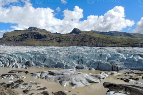 Gletsjer met ijsformaties en smeltwaterpoelen, berglandschap op de achtergrond onder een blauwe lucht.