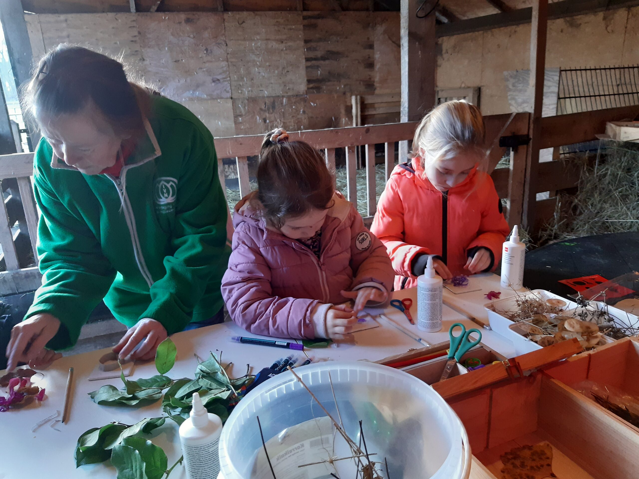 Drie mensen maken natuurknutsels aan een tafel met bladeren, lijm, en scharen in een schuur.