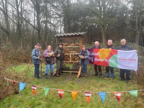 Groep mensen poseert met "NLdoet"-vlag bij houtopslag in bosrijke omgeving.