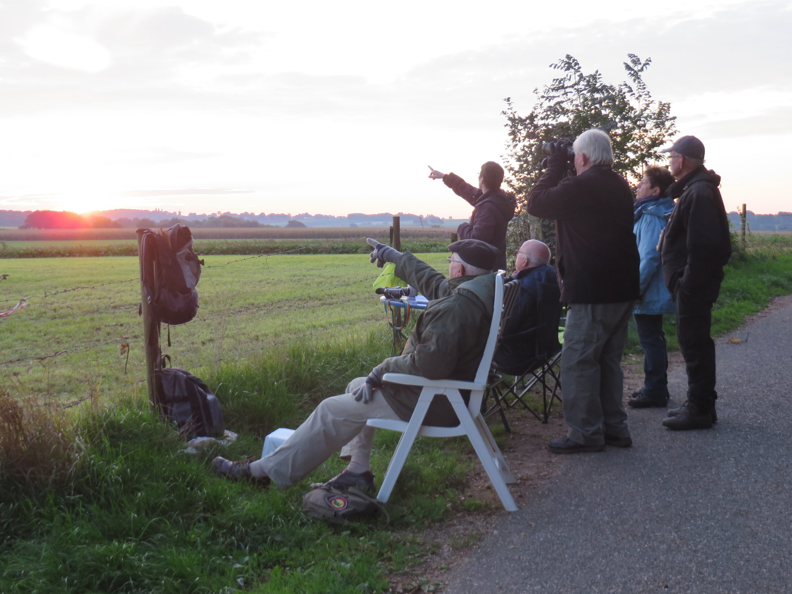 Mensen observeren de zonsondergang in een open landschap, sommigen met verrekijkers.