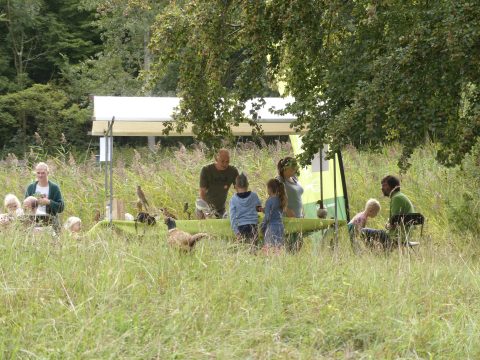Groep mensen onder luifel in grasveld, omgeven door bomen; informele bijeenkomst.