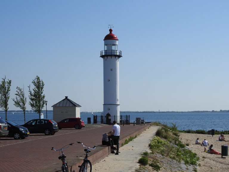 Vuurtoren bij de zee, met geparkeerde auto's, fietsen en mensen zittend op het strand.