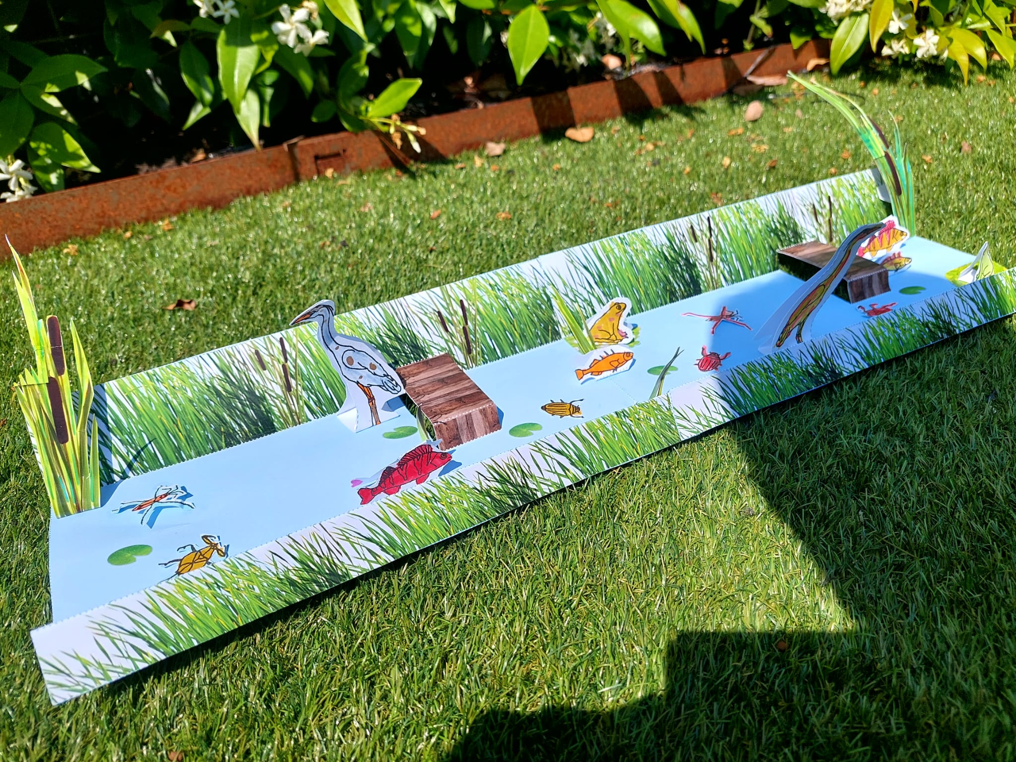 Papieren platte speelset van een rivieroever met dieren, op gras met groene planten op de achtergrond.