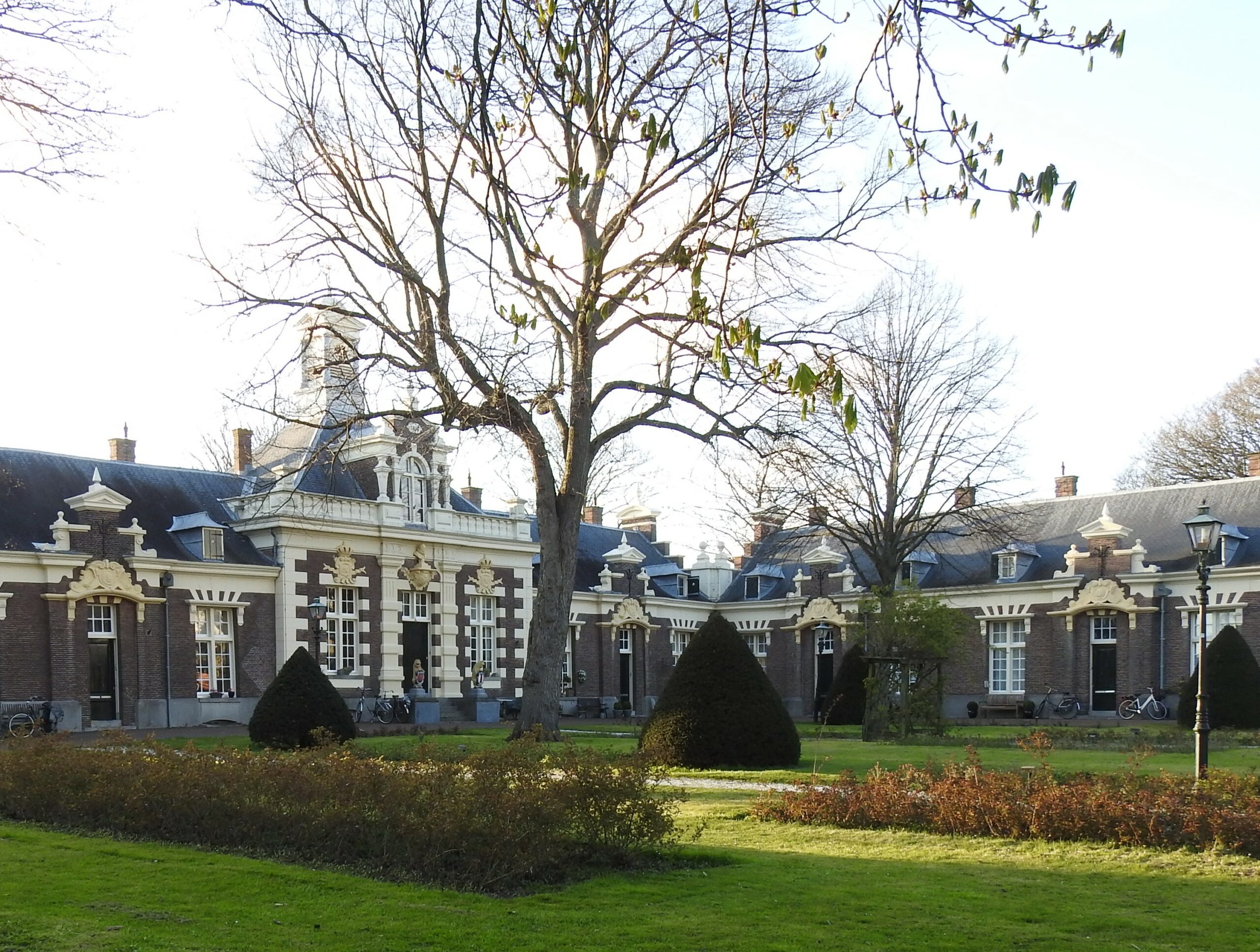 Historisch gebouw met rode bakstenen en witte details, omgeven door een tuin met kale bomen.