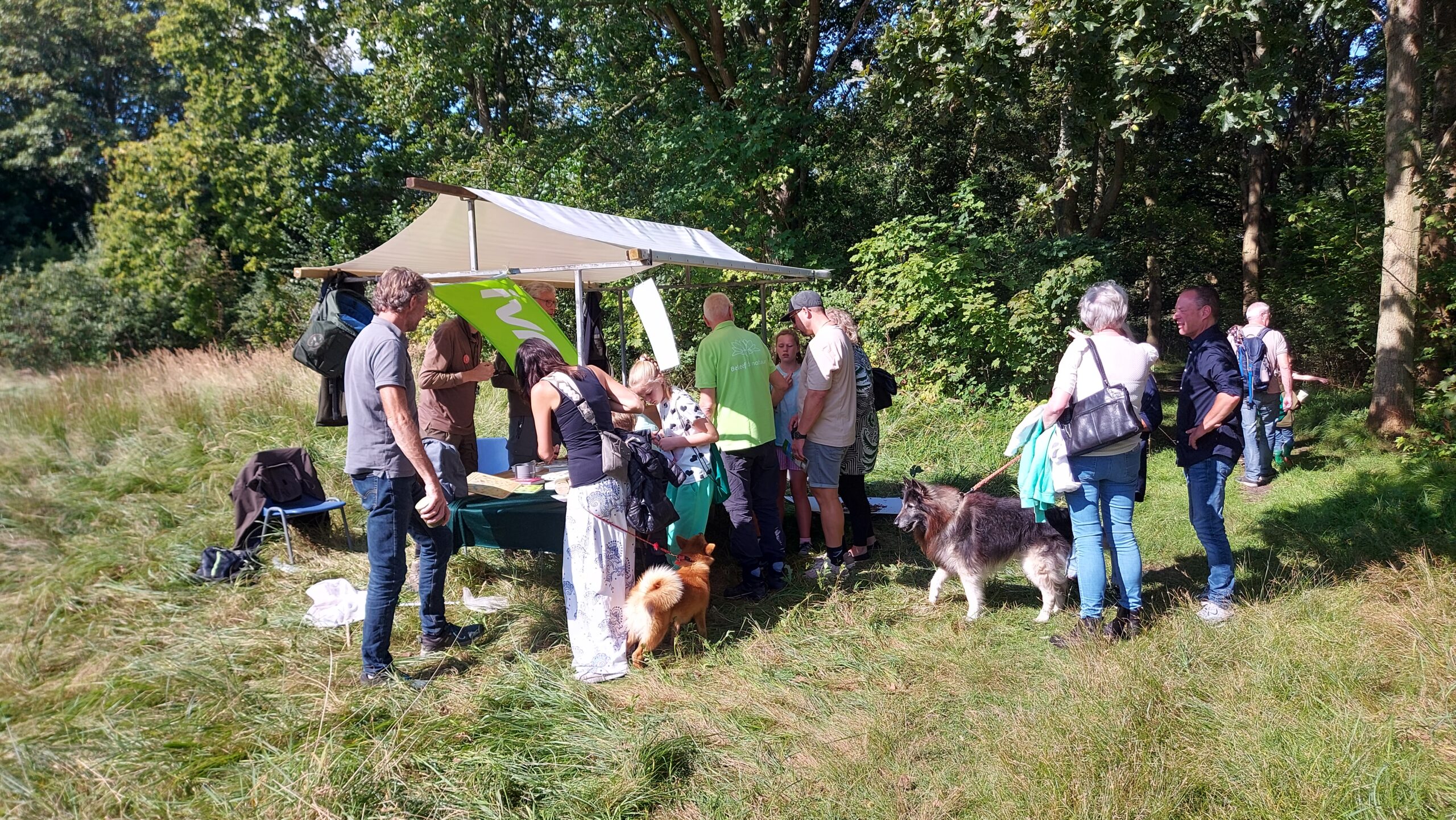 Mensen verzamelen zich rond een informatietent in een bosrijke omgeving, met honden aan de lijn.