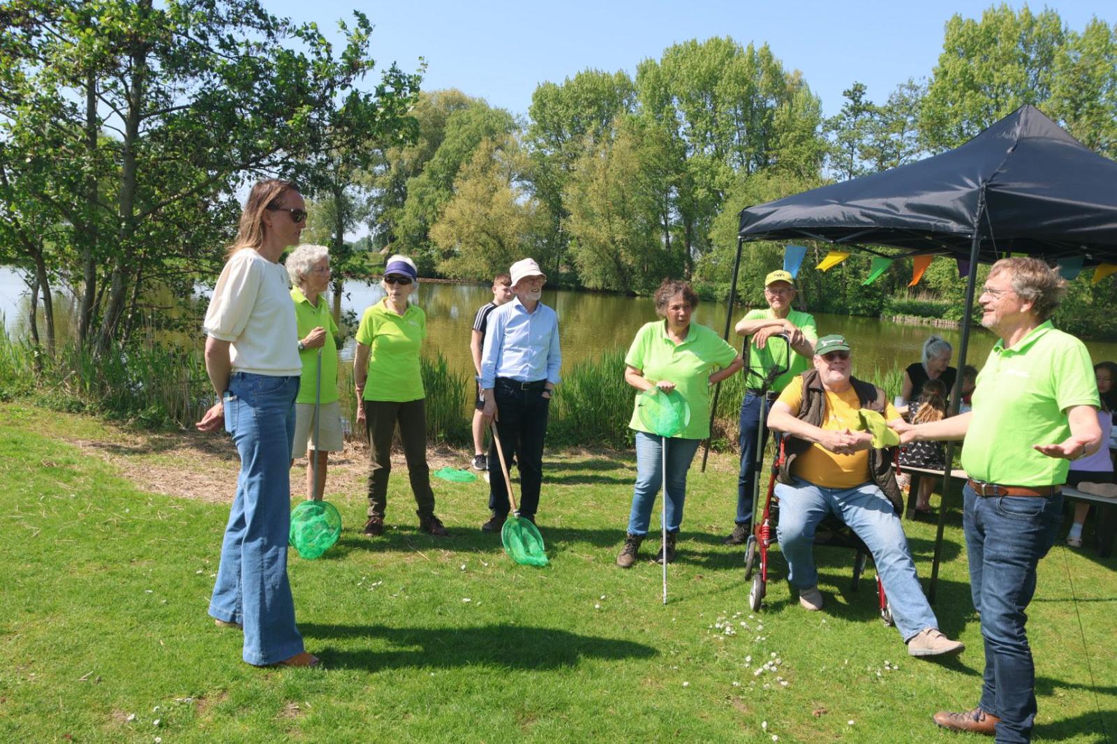 Groep mensen bij een meer met visnetten en groene shirts, onder een tent, omgeven door bomen.