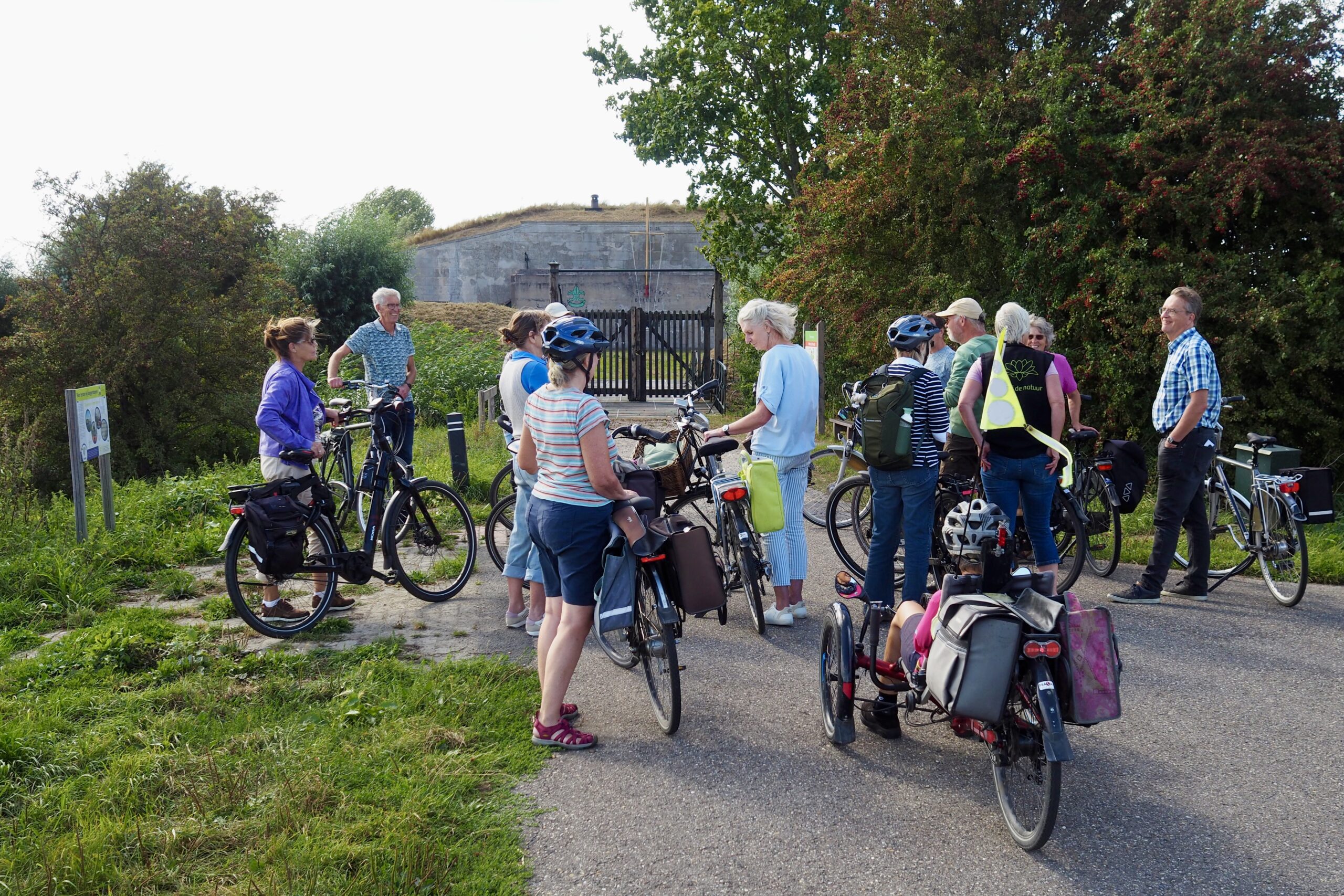 Groep mensen met fietsen, verzameld op een pad bij een fort, omgeven door groen en bomen.