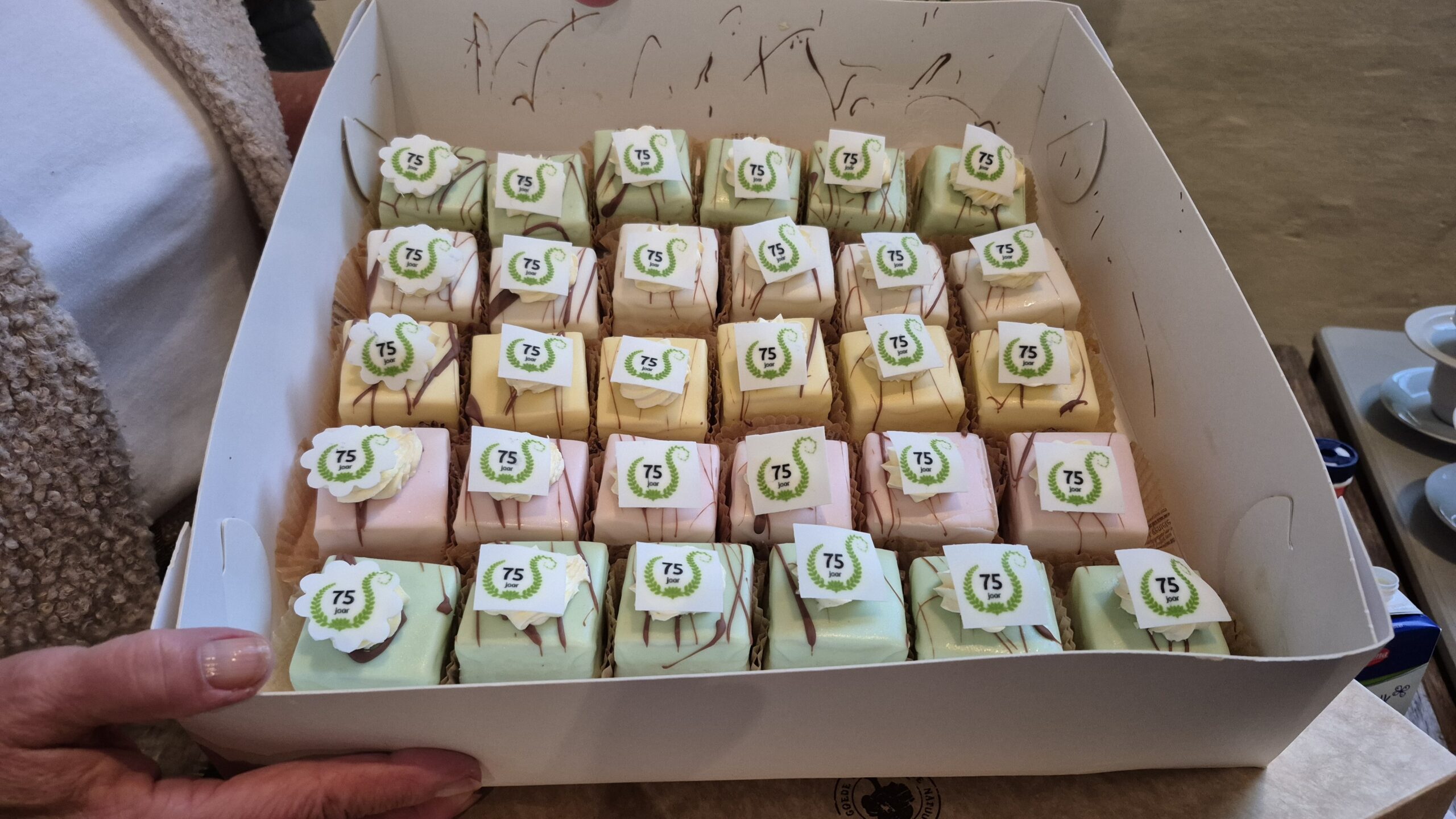 Een doos vol gebakjes met "75 jaar" logo en verschillende kleuren glazuur.