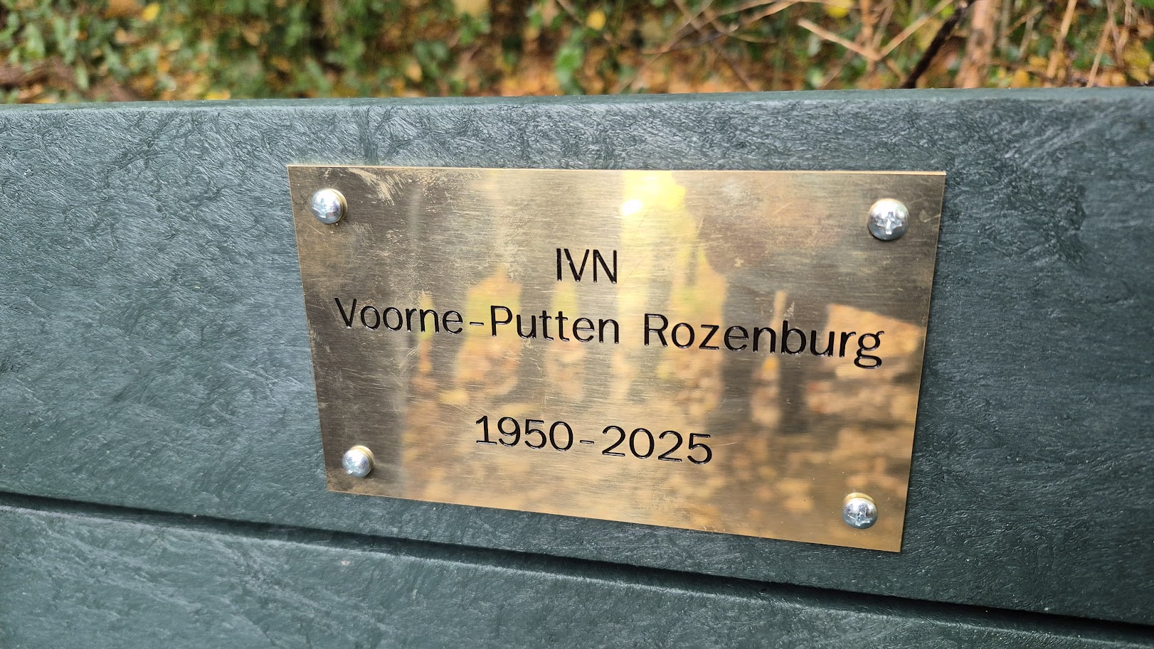 Messing plaquette op bankje met tekst "IVN Voorne-Putten Rozenburg 1950-2025", tegen groene achtergrond.