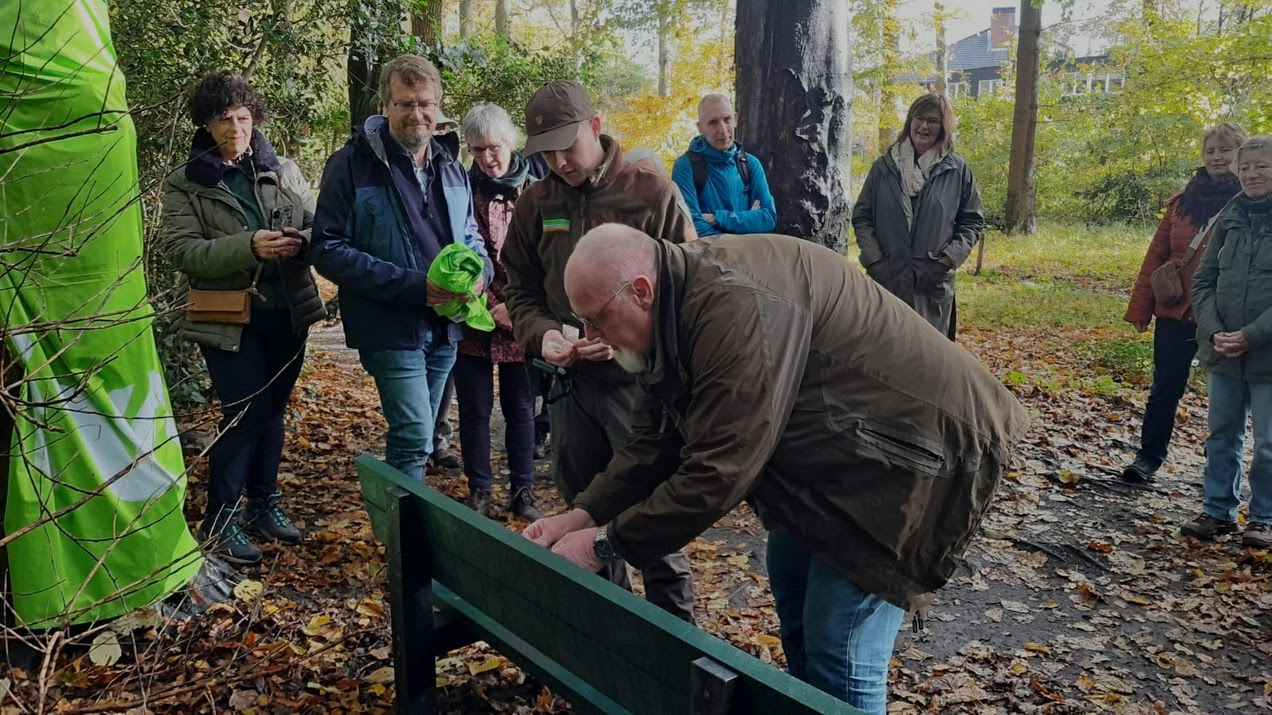 Groep mensen verzameld rond een bankje in een bos tijdens een ceremonie of onthulling.